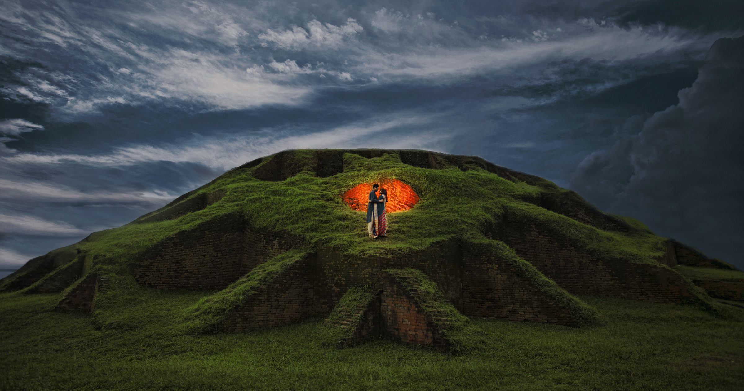 Bride and groom embracing inside a glowing brick archway atop a grass-covered hill at dusk